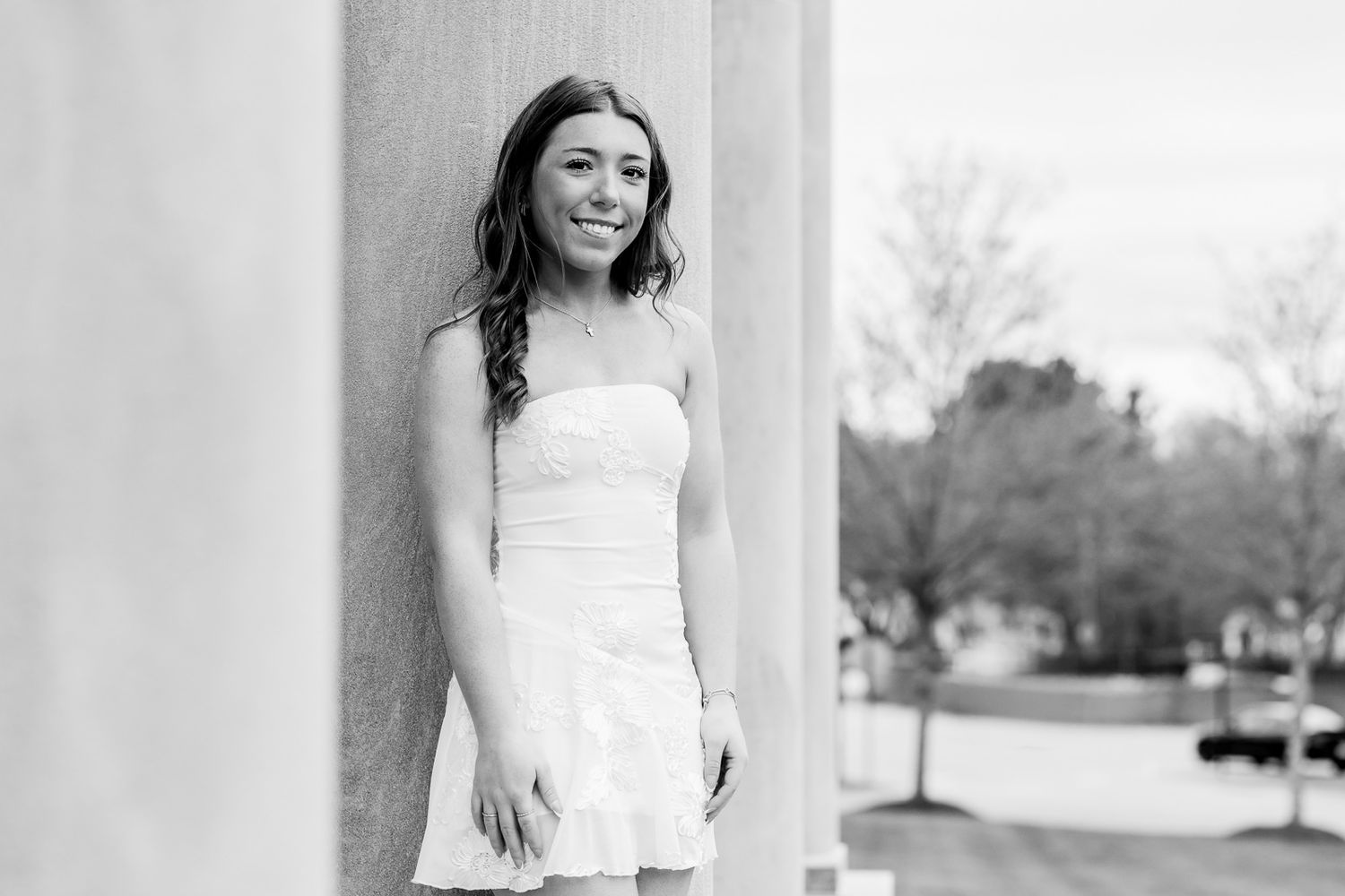 Young woman in white strapless dress smiling between columns in black and white portrait photo.