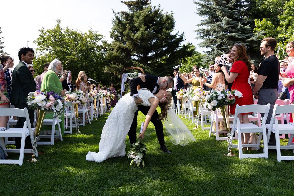 Bride and groom share a romantic dip kiss at outdoor wedding ceremony on green lawn with white chairs and guests.