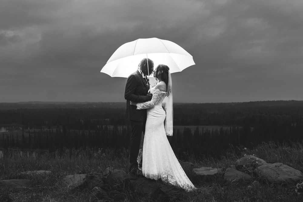 Black and white photo of a bride and groom kissing under a white umbrella on a cloudy wedding day.