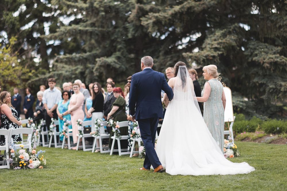 Bride walks down outdoor wedding aisle with guests seated on white chairs surrounded by lush green trees.