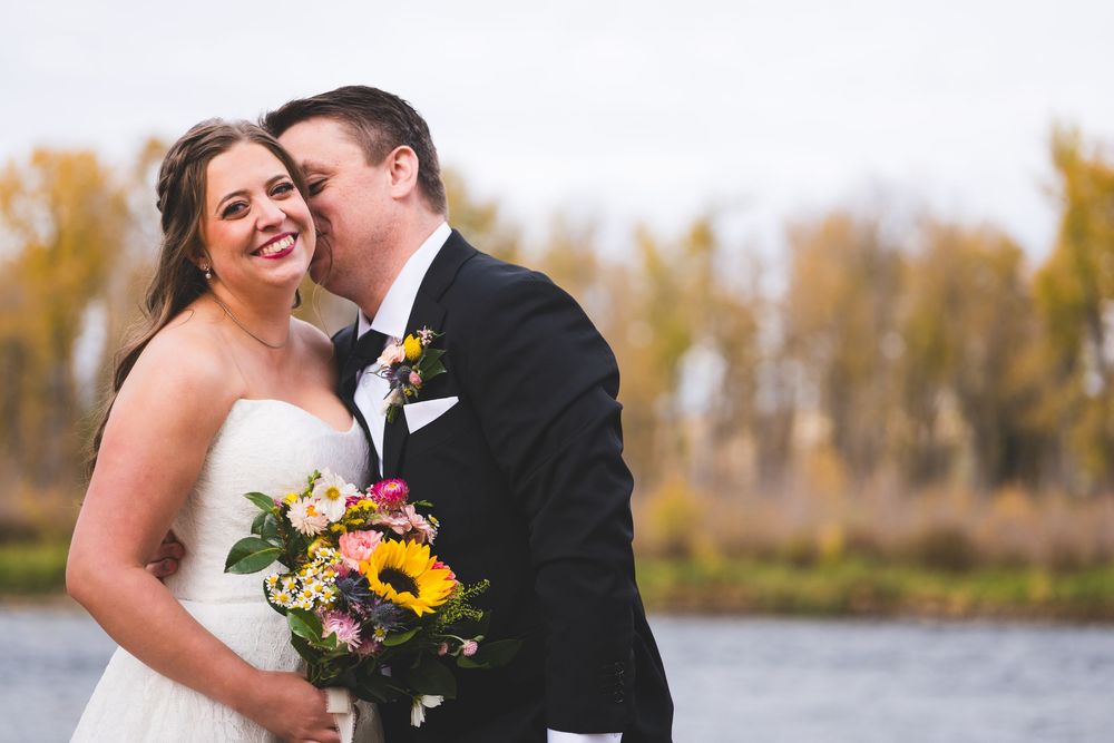 Bride and groom share a romantic kiss by a lake in autumn, bride holding a colorful sunflower bouquet.