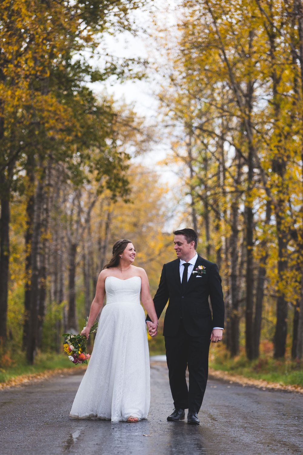 Bride and groom walk hand-in-hand down a rain-soaked road lined with golden autumn trees.