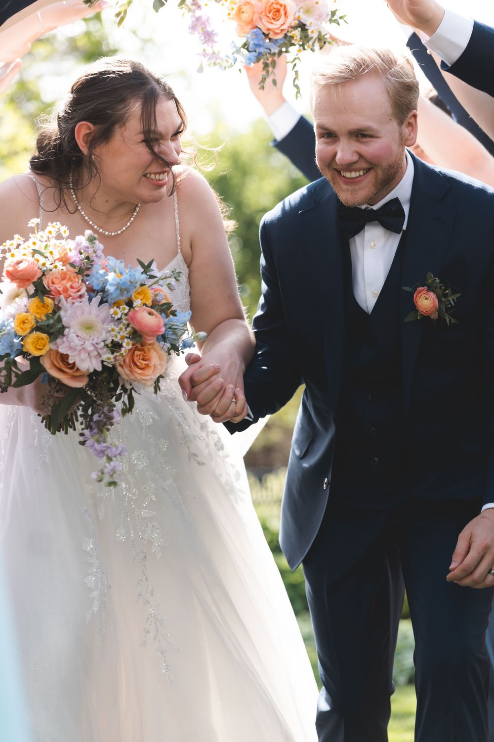 Joyful bride and groom holding hands, walking under floral arch, bride holding colorful bouquet, groom in navy suit.