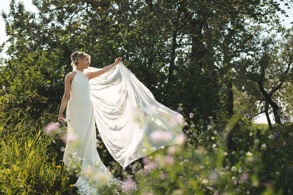 Bride in flowing white gown holding her veil in a lush garden surrounded by green trees and wildflowers.