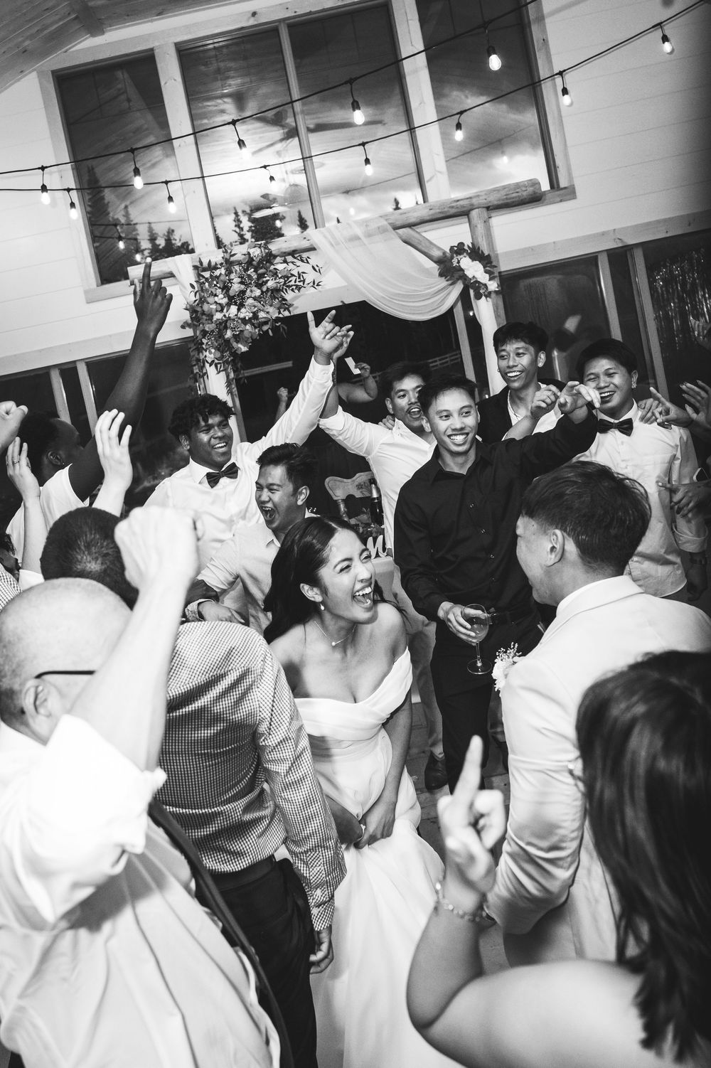 Black and white photo of a joyful bride dancing with guests at a lively wedding reception under string lights.