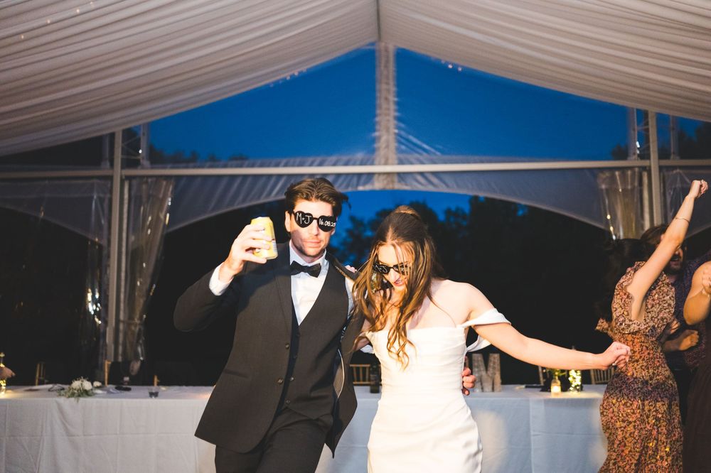Bride and groom dancing joyfully in sunglasses at outdoor wedding reception under white tent at dusk.