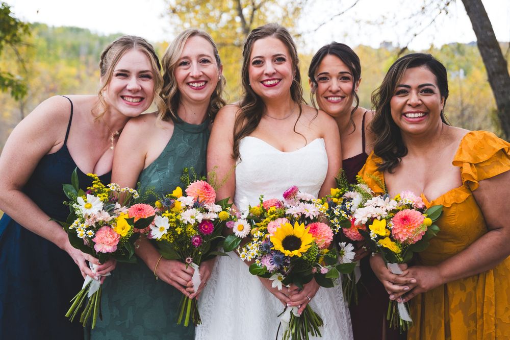 Bride in white gown poses with four bridesmaids in colorful dresses holding vibrant floral bouquets outdoors.