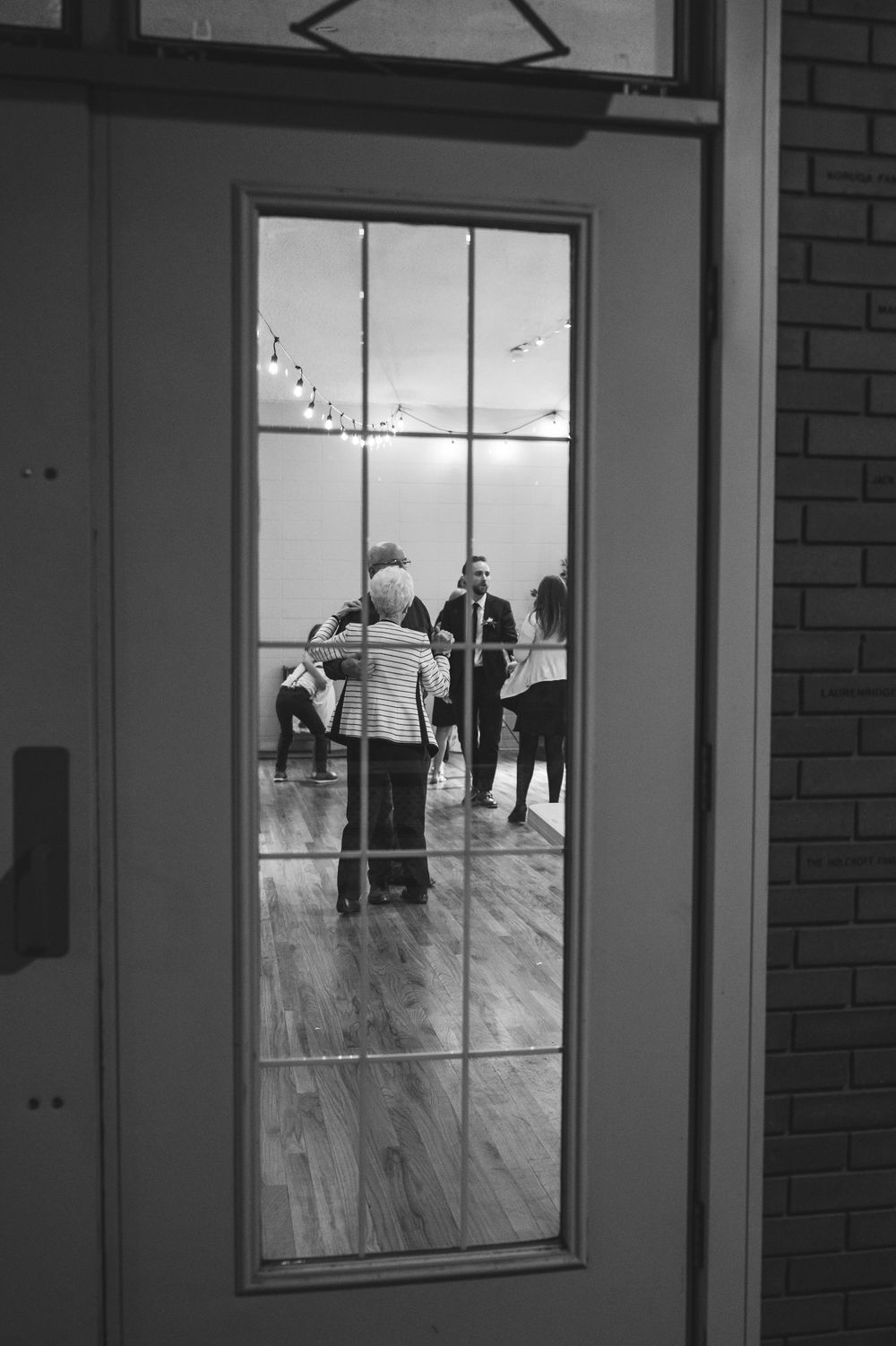 Black and white photo of people socializing inside a room viewed through a glass-paned door.