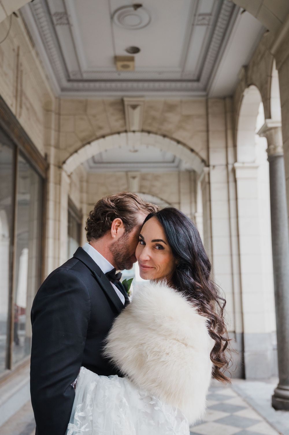 Bride in white fur stole and groom in black tuxedo pose in elegant arched hallway for wedding portrait.