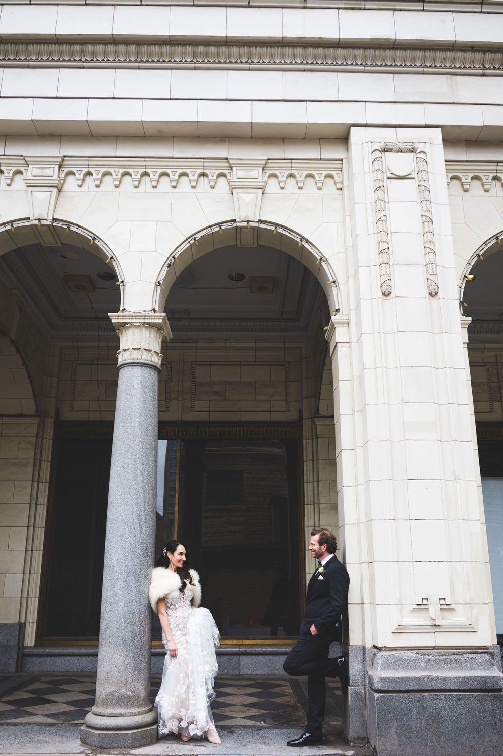 Bride and groom posing beside ornate stone columns and arched doorways of a classic neoclassical building.