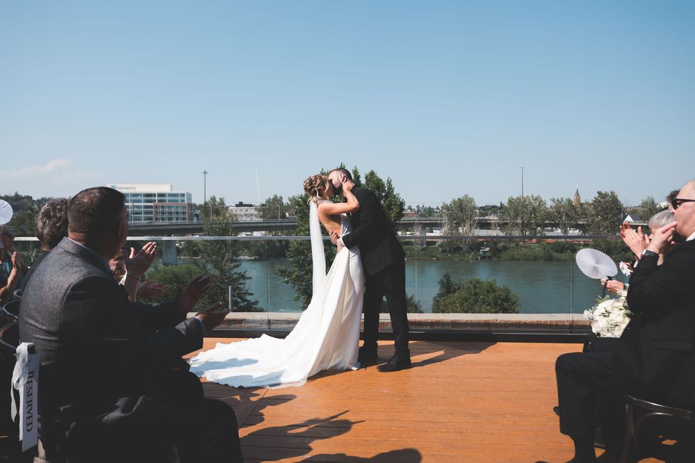 Bride and groom share first kiss at outdoor rooftop wedding ceremony with river and city skyline backdrop.