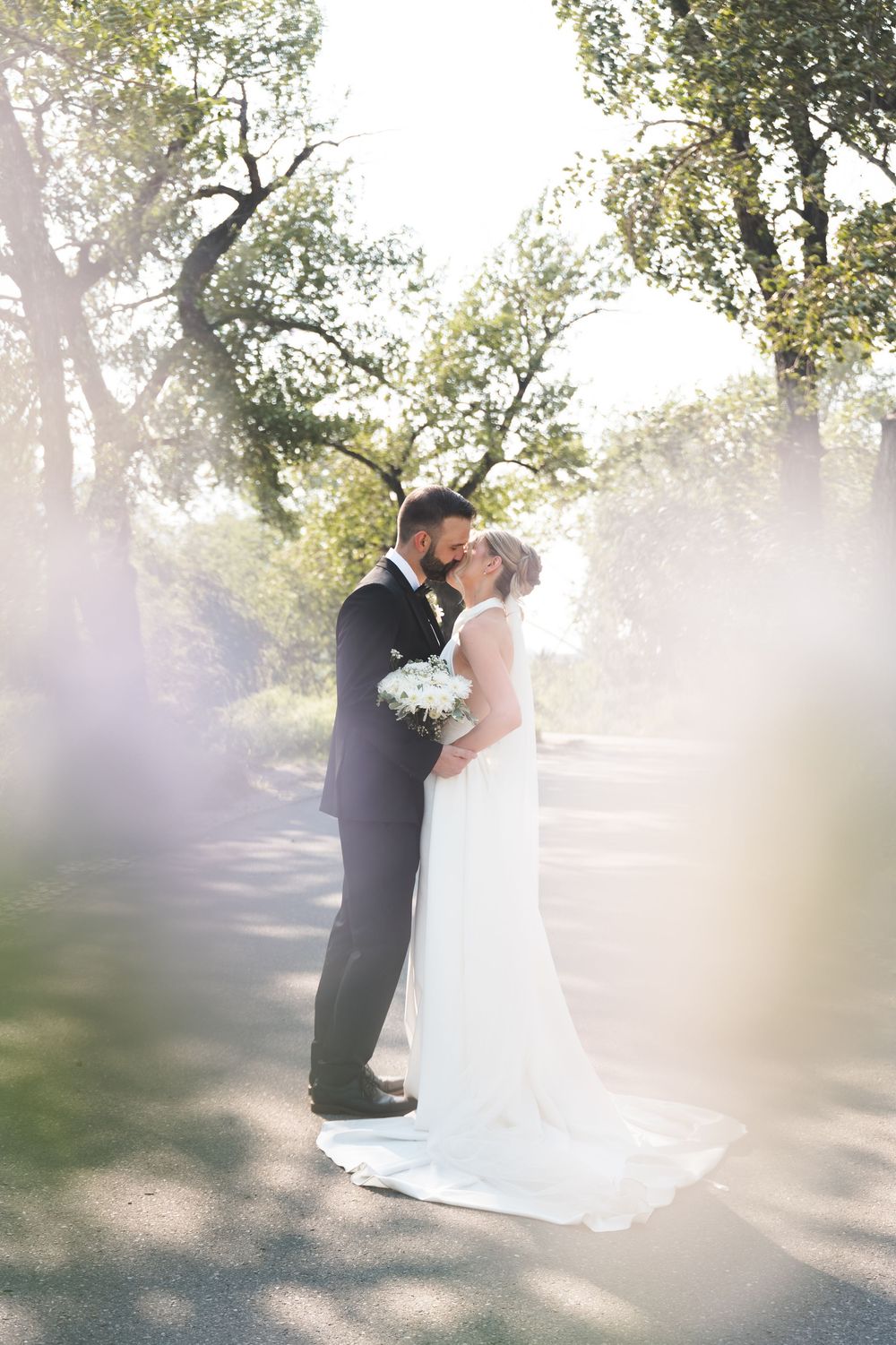 Bride and groom share a romantic kiss outdoors, surrounded by lush green trees, bride holding white bouquet in elegant wedding dress.