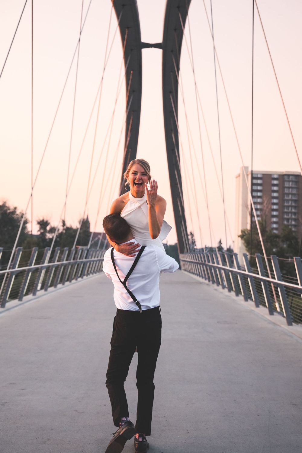 A joyful couple on a suspension bridge, man lifting woman in white dress at sunset.