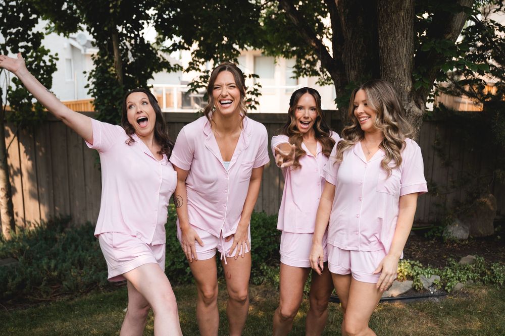 Four women laughing and posing outdoors in matching pink pajama sets during a fun bridal party photoshoot.