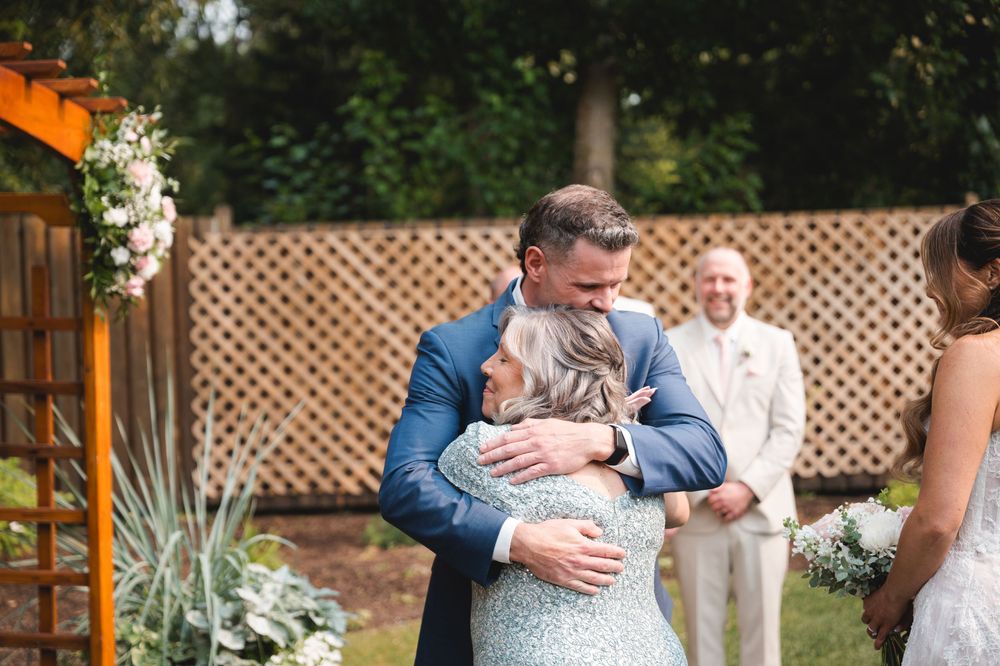 Groom in blue suit hugs a guest at an outdoor wedding ceremony with lattice backdrop and floral arch.