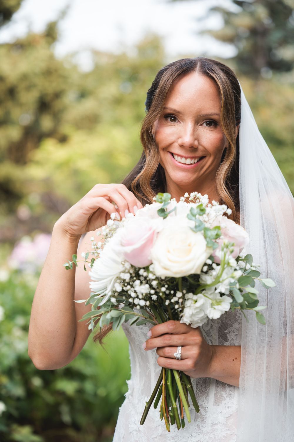 Smiling bride in white lace dress and veil holds pink and white rose bouquet outdoors.
