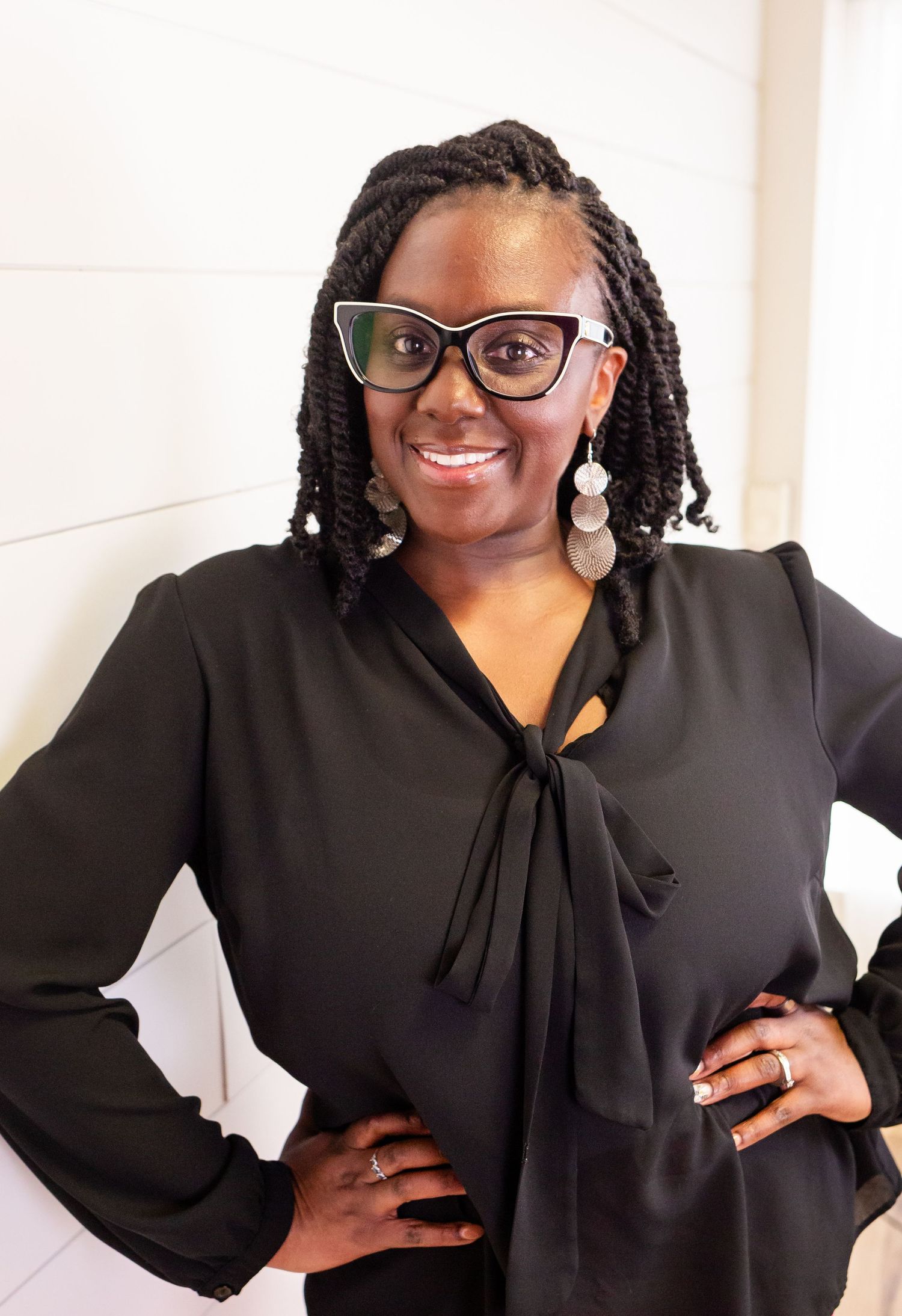 Smiling woman with braids, glasses, and black tie-front blouse posing with hands on hips.