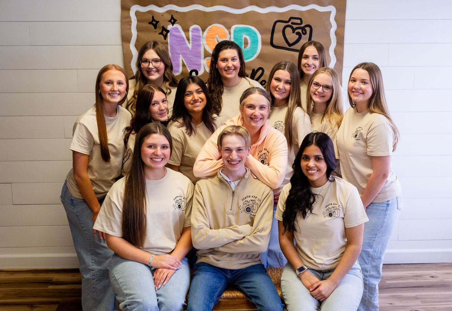 Group of young adults in matching beige shirts posing together indoors in front of a colorful NOR sign.