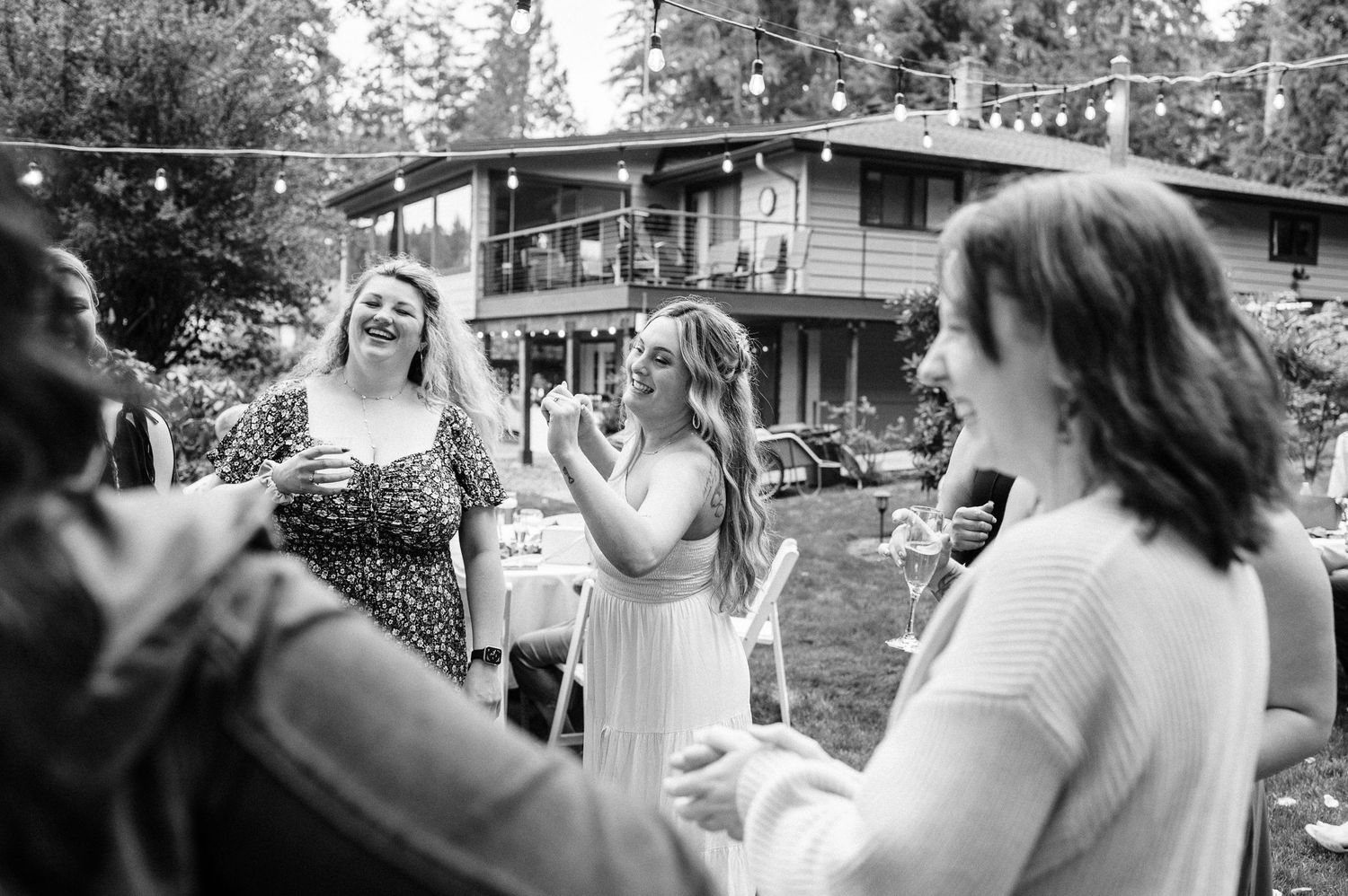 Women laughing and dancing at an outdoor backyard wedding with string lights and a house in background.