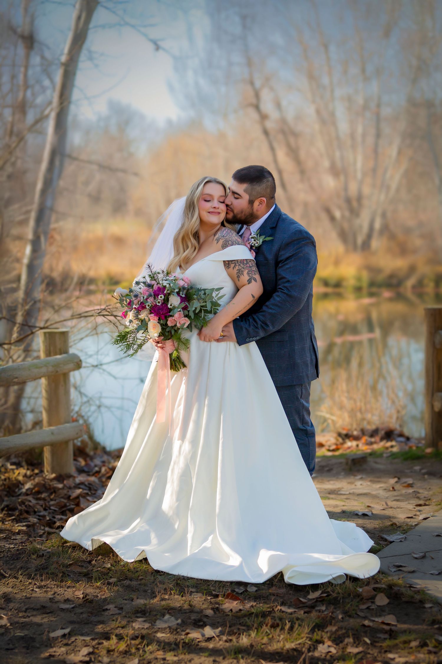 Bride and groom posing groom kissing bride on cheek in front of pond