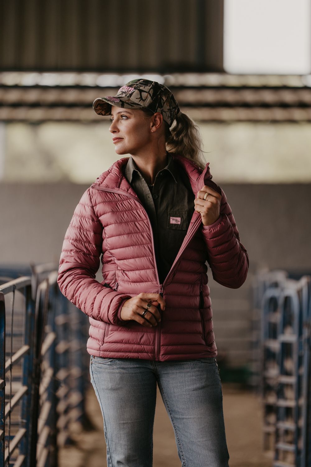 Woman in pink quilted puffer jacket and camo cap standing in a farm setting.