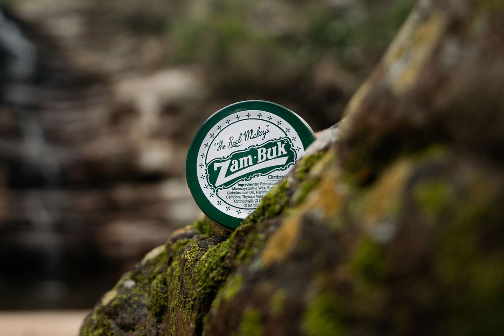 A small tin of Zam-Buk ointment rests on a mossy rock with a blurred natural background.