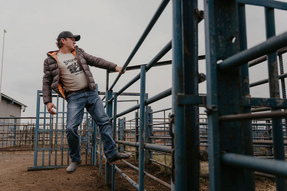 A man in a jacket and jeans leans on a metal livestock fence at a rural farm.