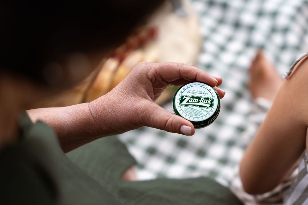 A person holds a small round tin of Zam-Buk ointment outdoors on a checkered blanket.