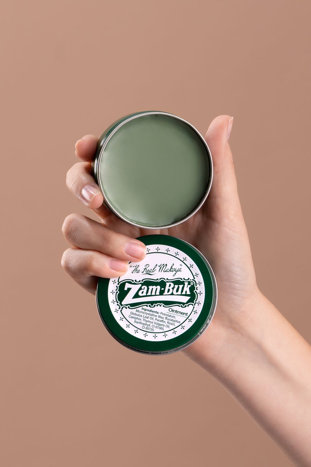 A hand holds an open tin of Zam-Buk herbal ointment against a neutral beige background.