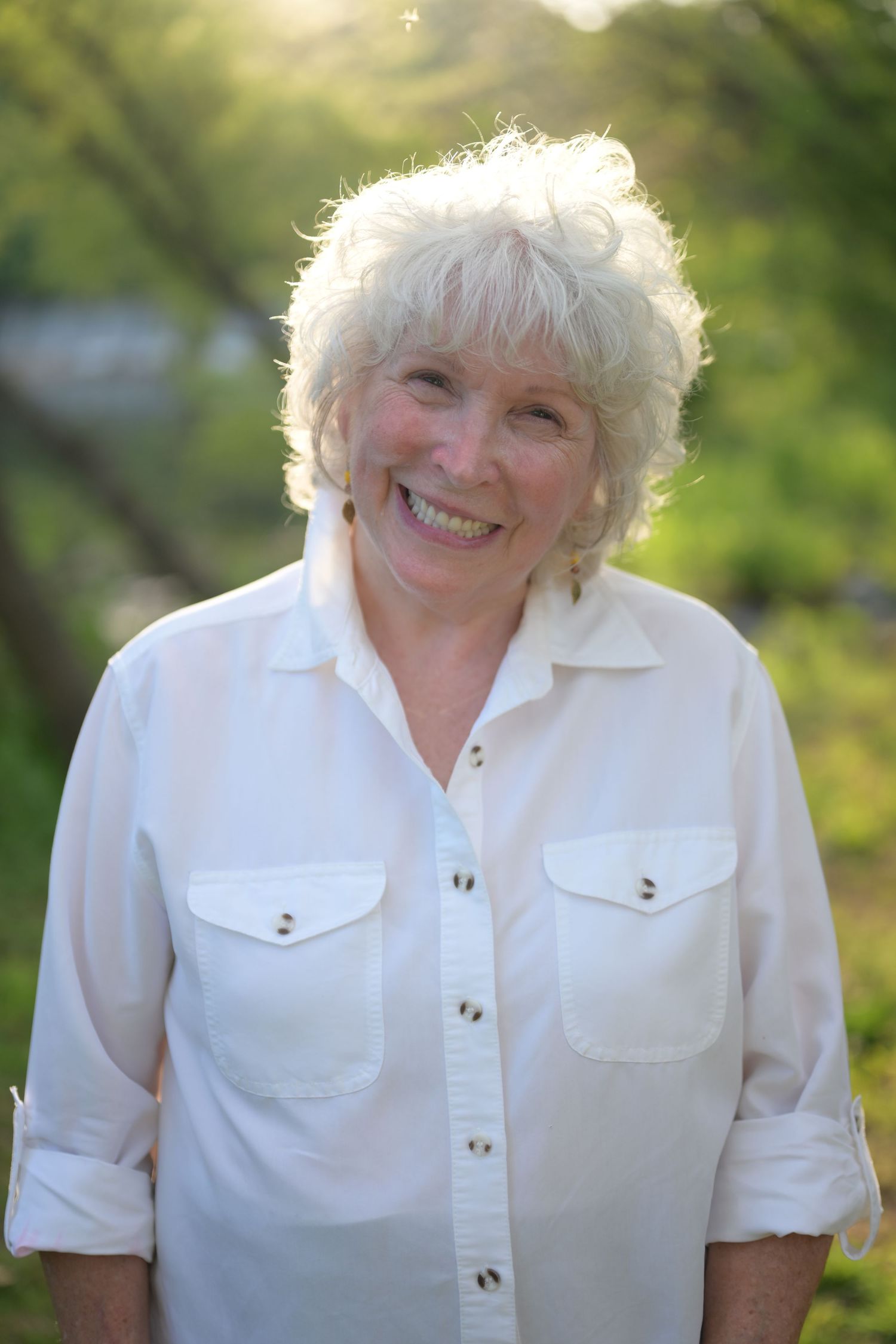 Smiling older woman with white curly hair wearing a white button-up shirt outdoors in natural green setting.