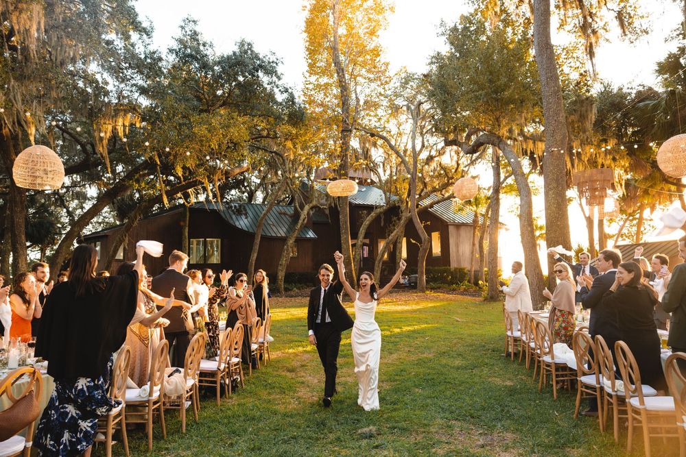 Newlyweds walk down outdoor aisle under oak trees at golden hour wedding ceremony with seated guests.