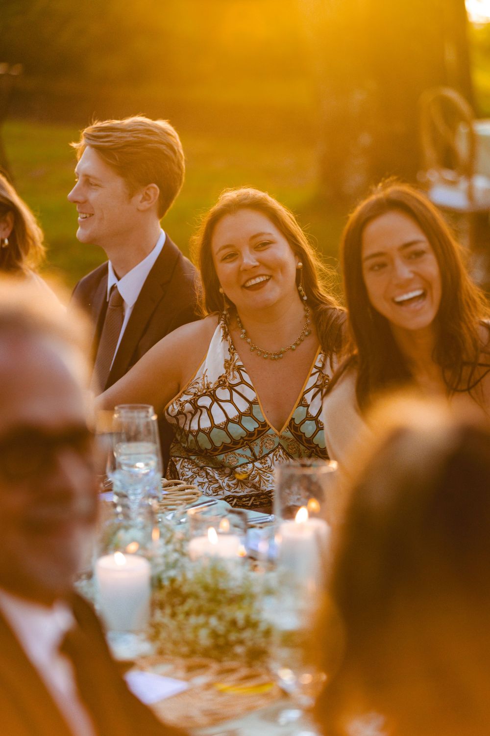 Wedding guests laugh and smile at candlelit outdoor reception table during golden hour celebration.
