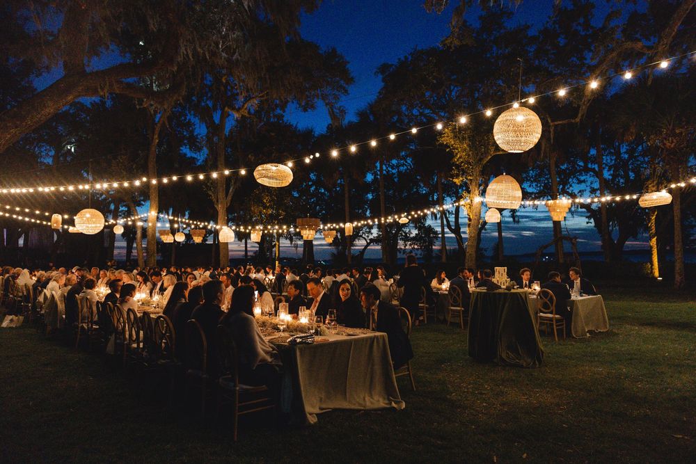 Elegant outdoor evening reception with string lights and paper lanterns illuminating guests dining under trees at night.