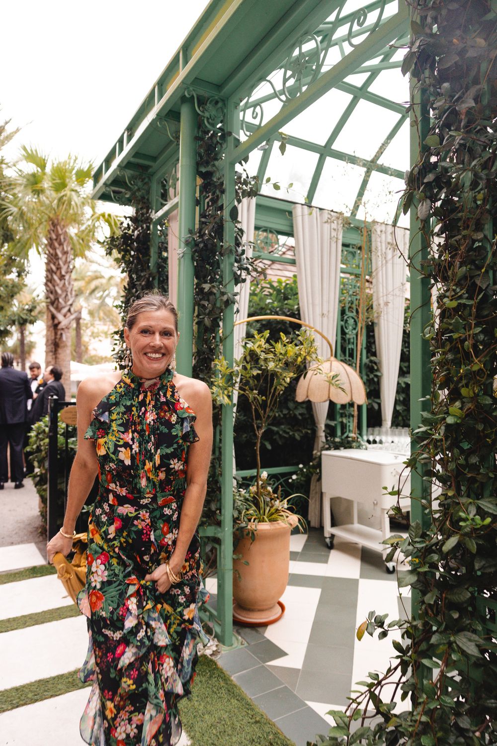 Woman in floral halter dress smiling in front of green greenhouse gazebo with tropical plants and white curtains.