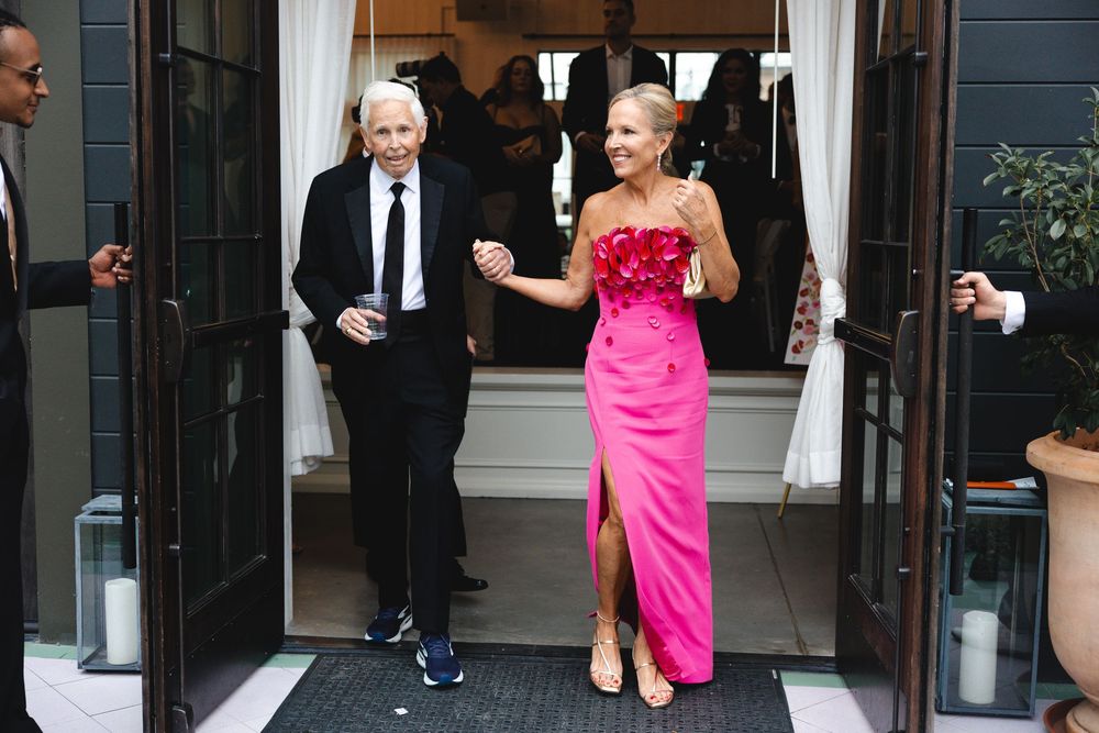Elegant couple in formal attire, man in black suit and woman in pink gown, walking through doorway at event.