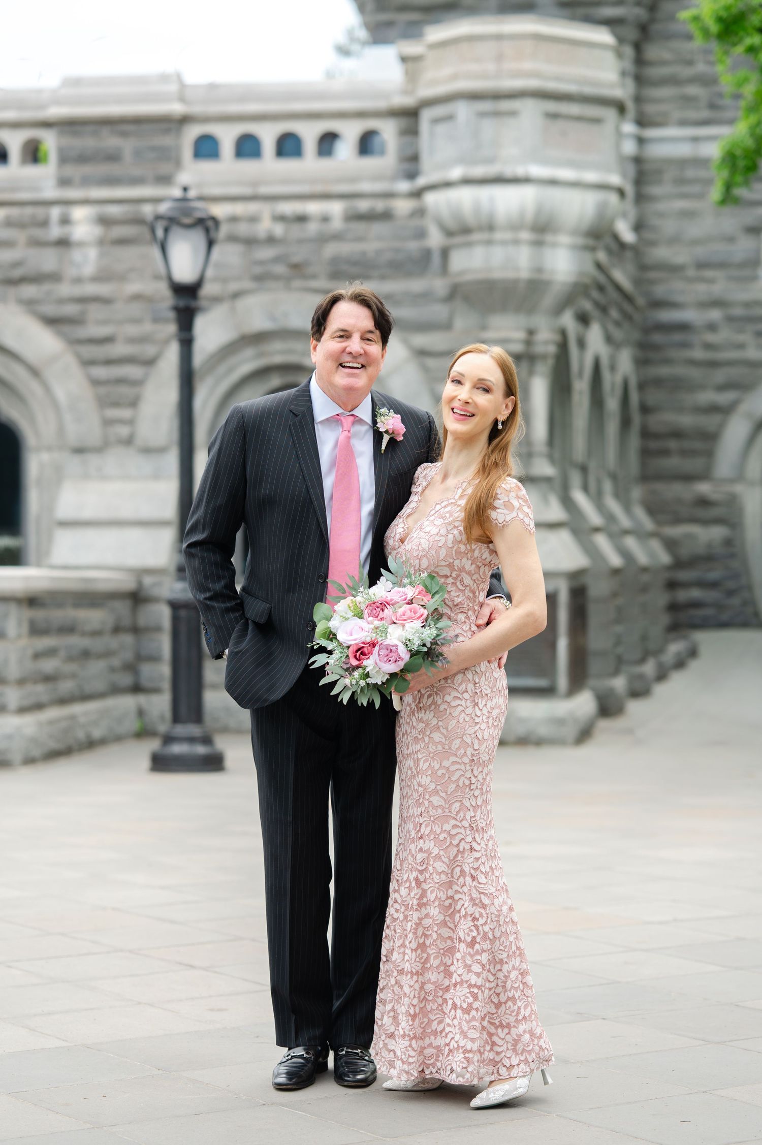 Couple posing in wedding attire outside a stone castle, woman in pink lace gown holding bouquet, man in dark suit.