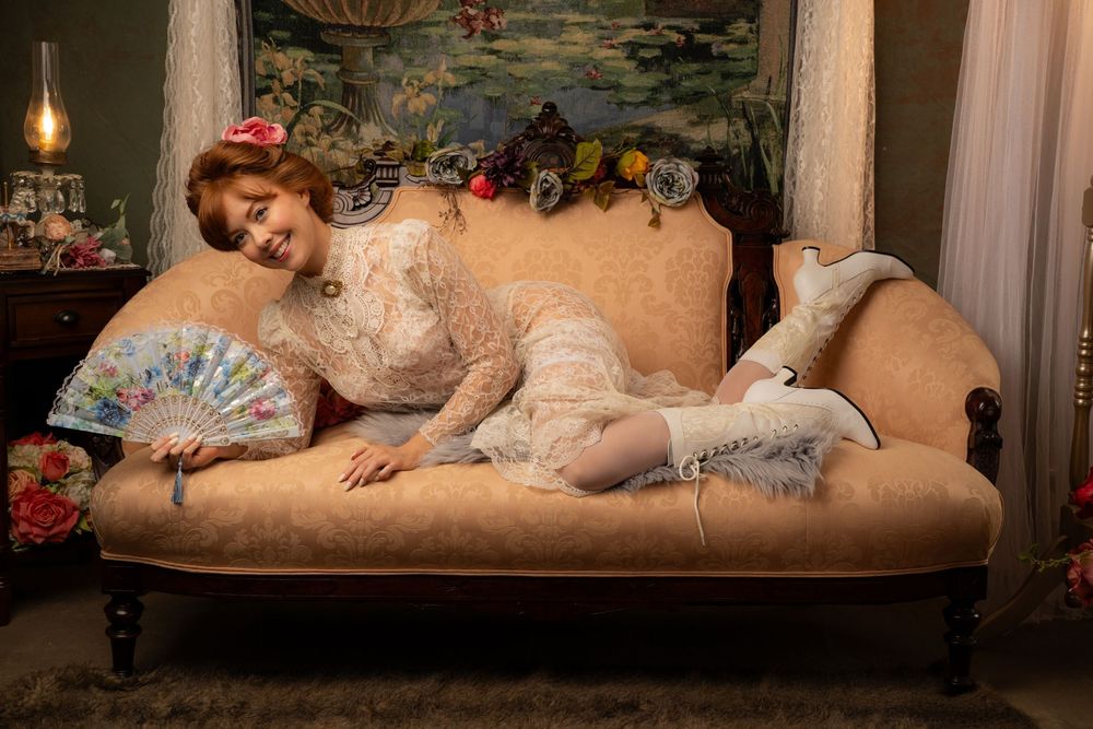 Woman in vintage lace dress reclines on antique chaise lounge holding decorative fan in Victorian-style room.