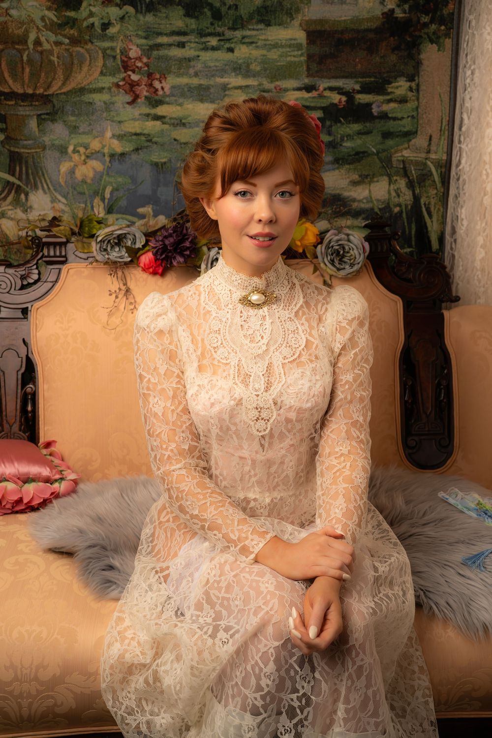 A young woman in a white lace Victorian dress sits on a vintage sofa before a floral tapestry backdrop.