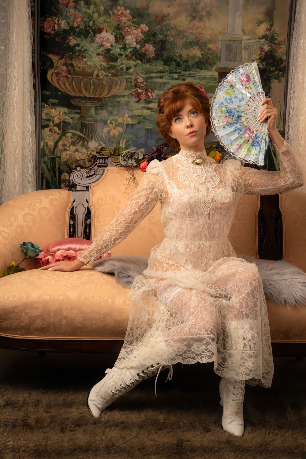 Elegant redhead in vintage white lace gown raising decorative fan while seated on antique chaise lounge.