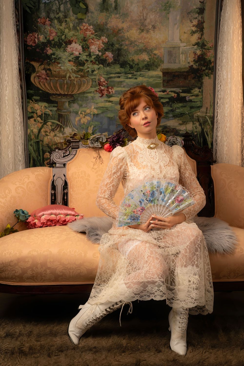 Victorian woman in white lace dress holding floral fan seated on ornate sofa before floral tapestry.