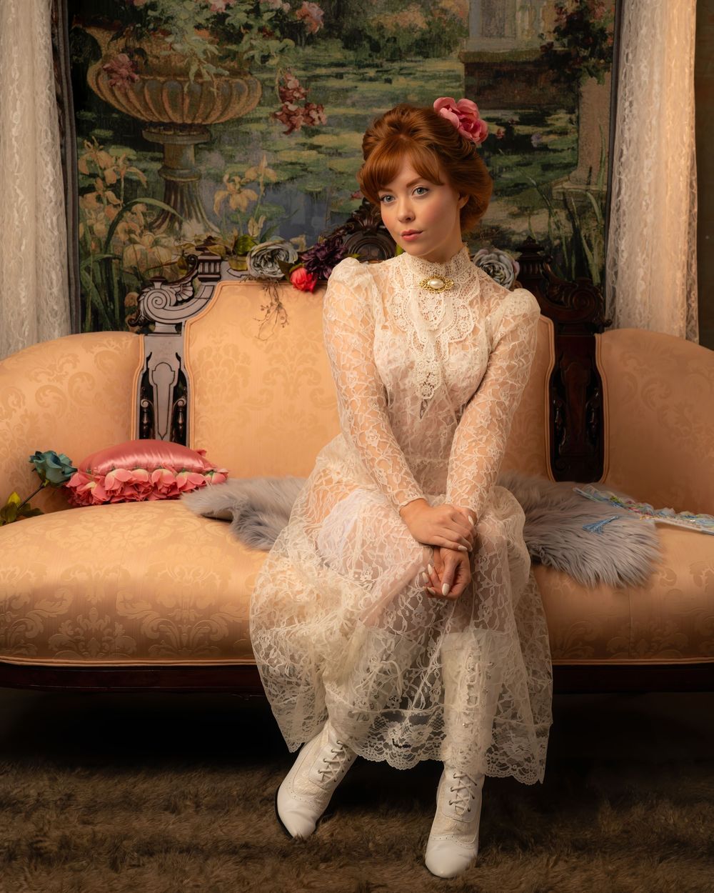 Woman in vintage white lace dress and boots sits on ornate sofa before floral tapestry.