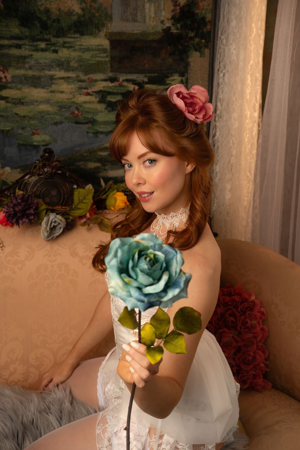 Red-haired woman in white lace holding a blue rose, sitting on a beige sofa with floral decor.