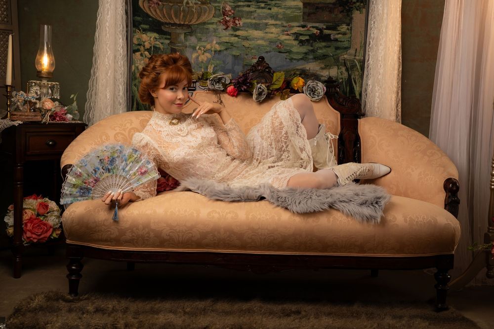 Woman in vintage white lace dress reclines on antique sofa holding a fan in an elegantly decorated Victorian-style room.