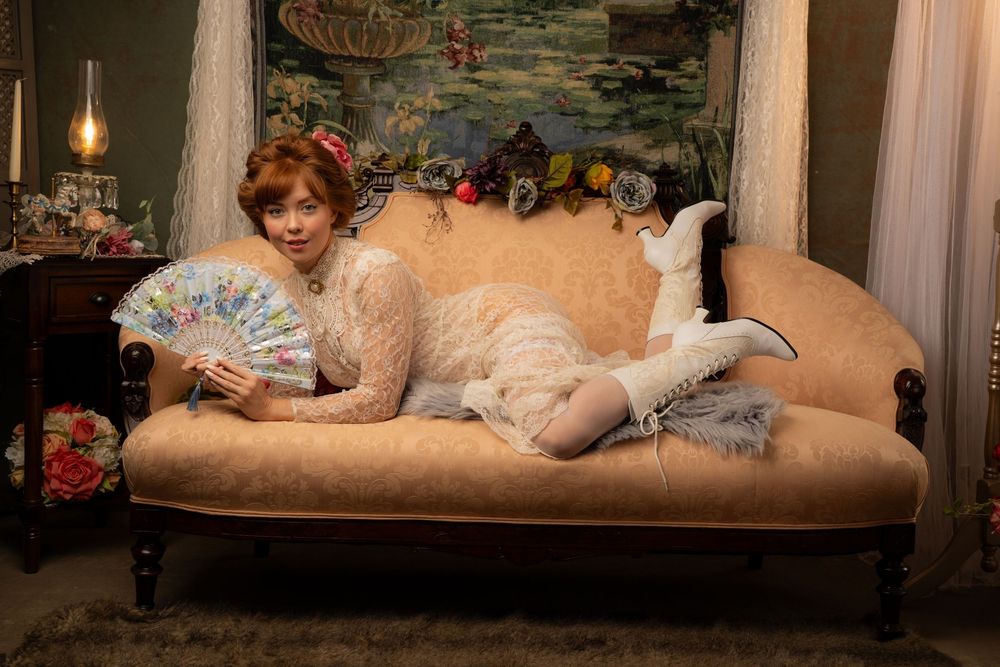 Woman in white lace dress holding decorative fan reclines on vintage sofa in elegant Victorian-style room.