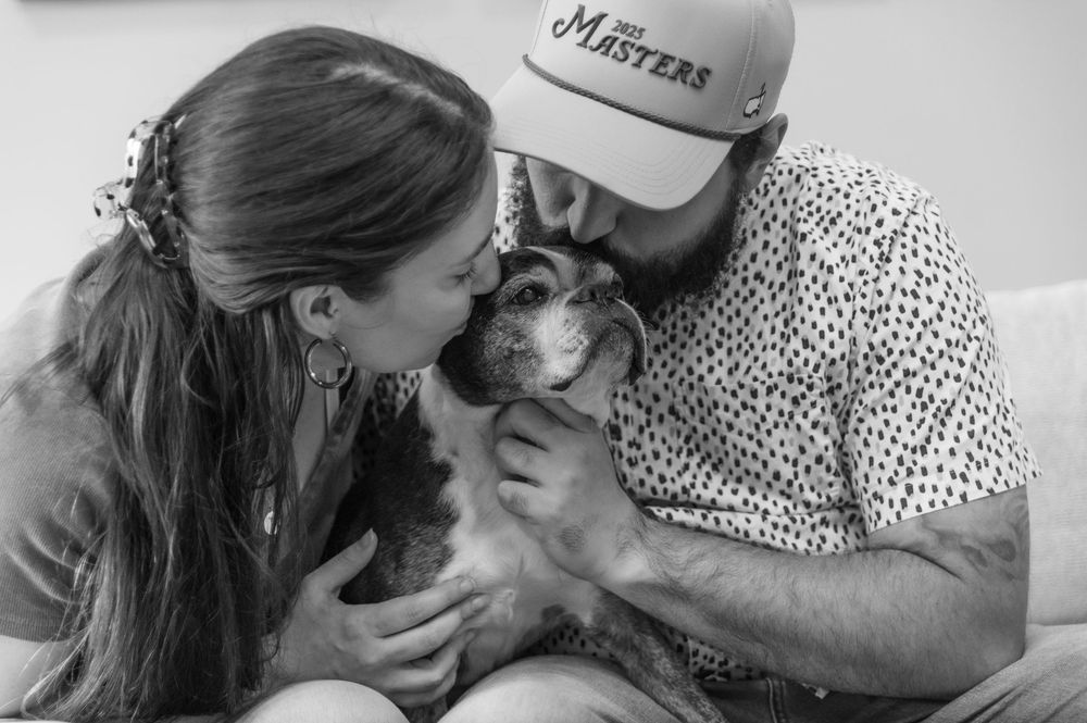 Black and white photo of a couple kissing their small dog on a couch.