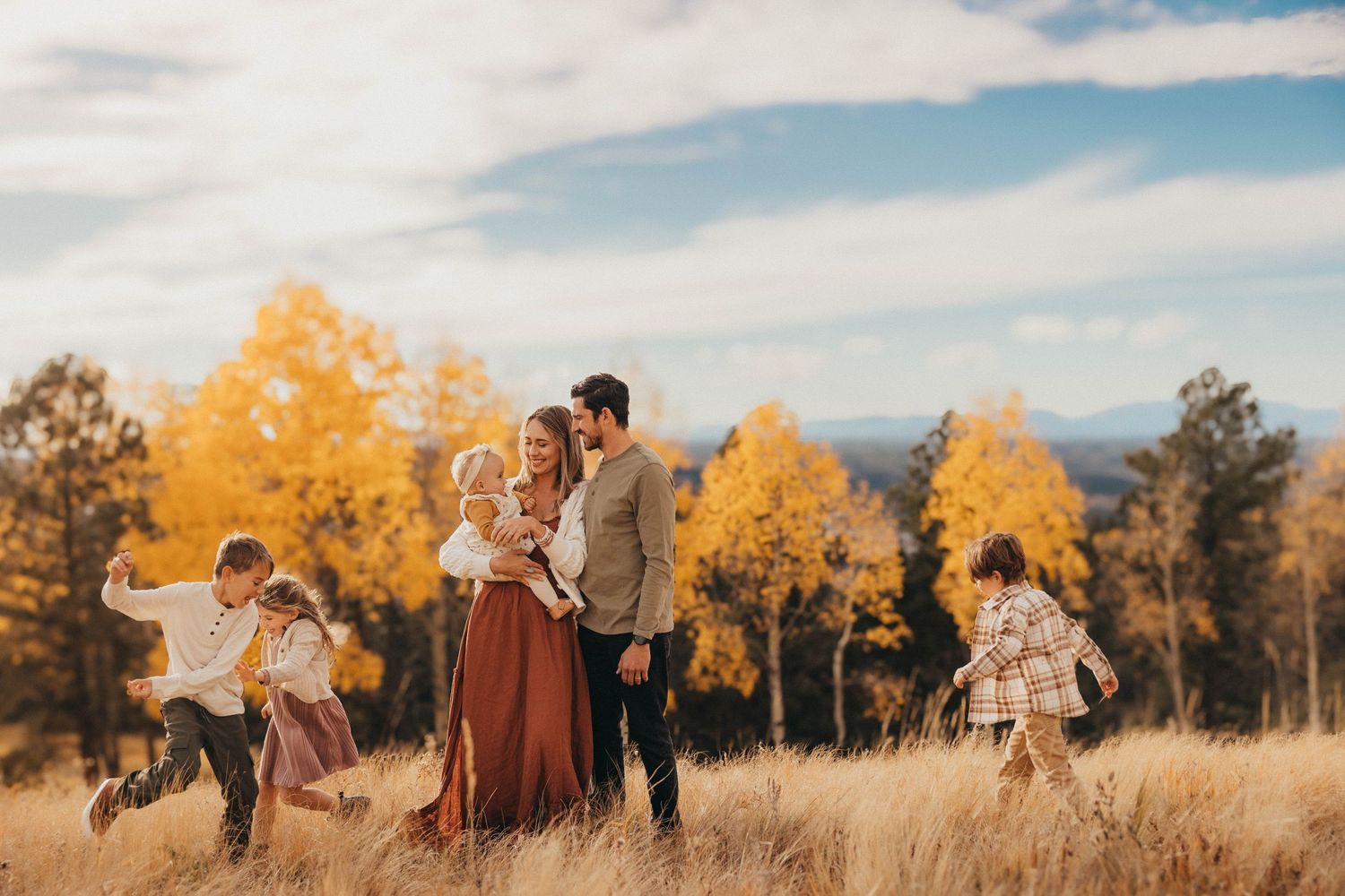 Family enjoying autumn walk in golden meadow with yellow aspen trees and mountains under cloudy sky.