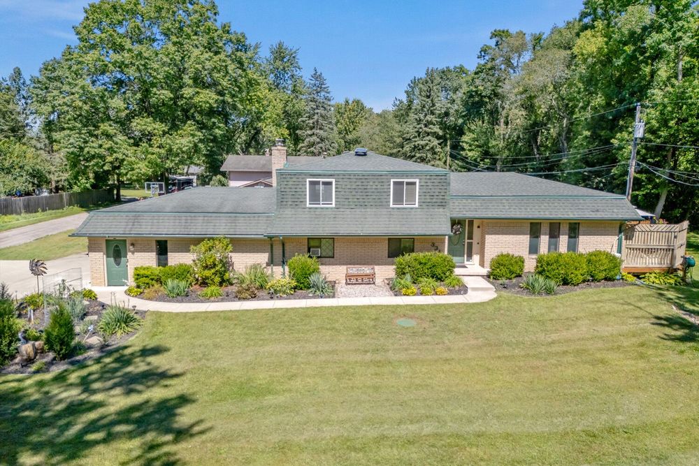 Brick and wood ranch home with wide front lawn, manicured landscaping, and tall trees in background.