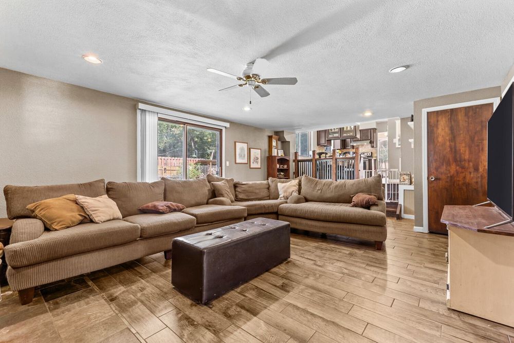Open living room with large brown sectional sofa, wood-look flooring, ceiling fan, and view into kitchen.