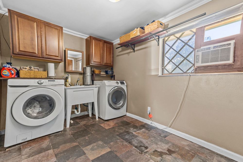 Laundry room with front-load washer and dryer, wooden cabinets, window AC unit, and stone tile floor.