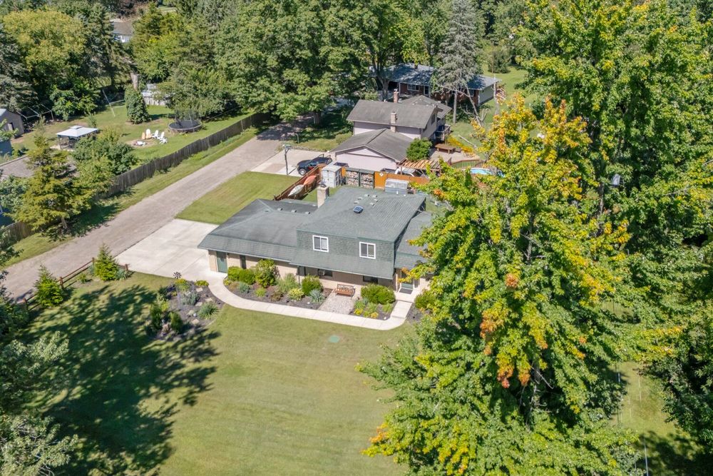 Aerial view of a ranch home with large driveway, green lawn, patio area, and dense tree surroundings.